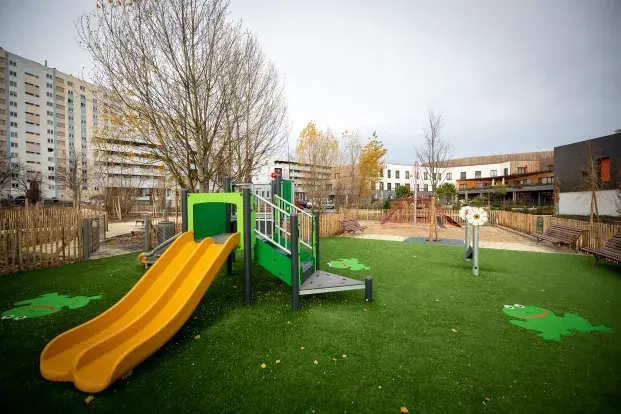 Une aire de jeux colorée avec un toboggan, du gazon synthétique et des immeubles en arrière-plan.