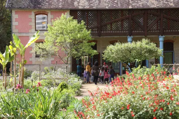 Un groupe d'enfants et d'adultes se rassemble devant une maison entourée d'un jardin, rempli de fleurs colorées et de plantes verdoyantes.