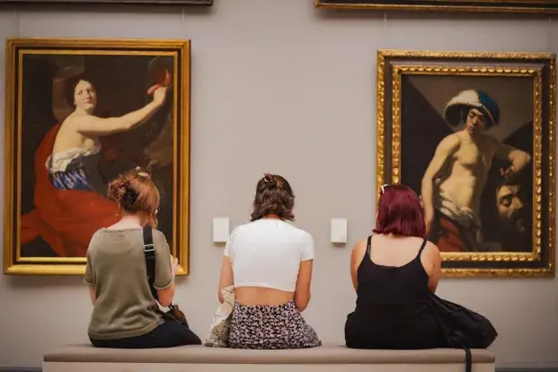 Trois femmes de dos assises sur un banc regardant deux tableaux.