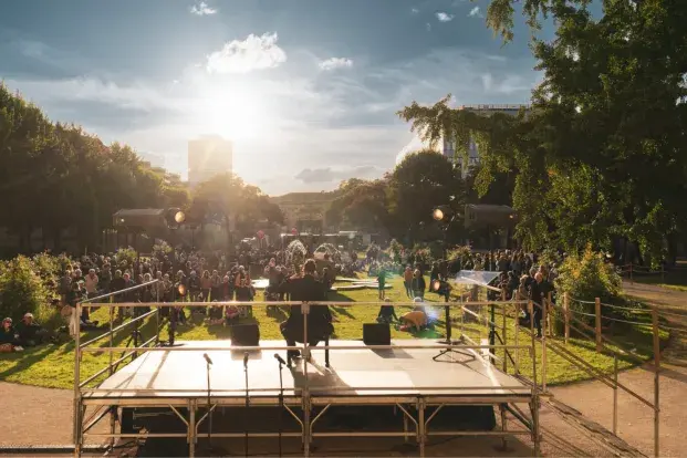 ue depuis l’arrière d’une scène installée en plein air, en fin de journée. Un artiste est assis sur une chaise au centre de la scène, face à une grande foule rassemblée dans un parc verdoyant, baigné par la lumière dorée du soleil couchant.