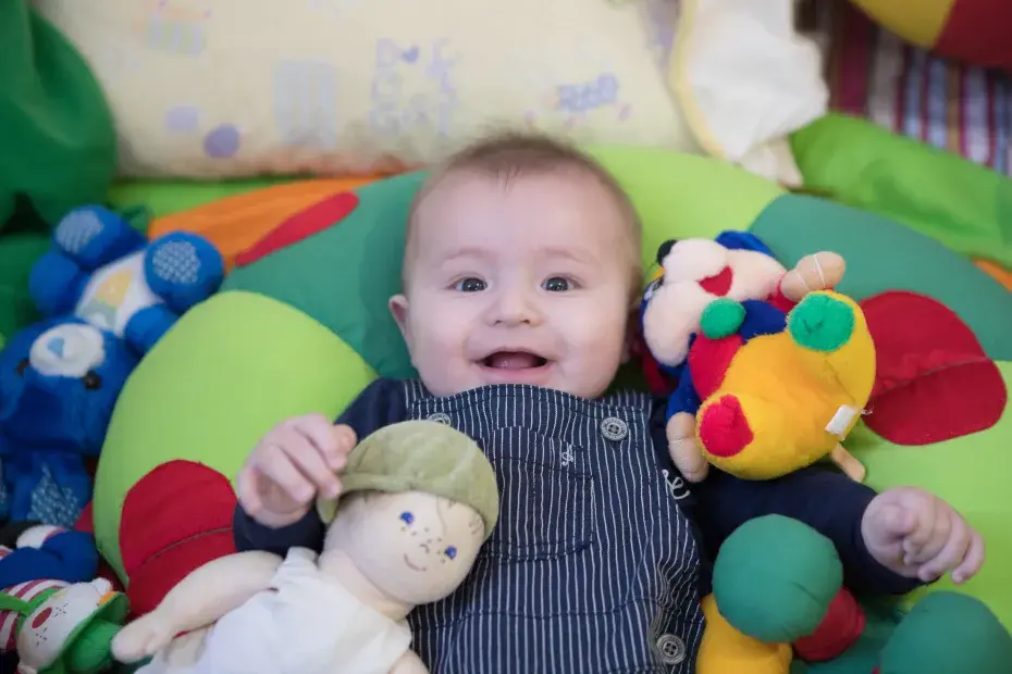 Un bébé souriant est bien installé dans des coussins entouré de doudous.