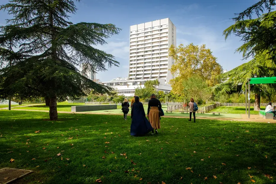 Des personnes marchent dans un parc avec de l’herbe et des arbres, et un grand immeuble blanc au fond.