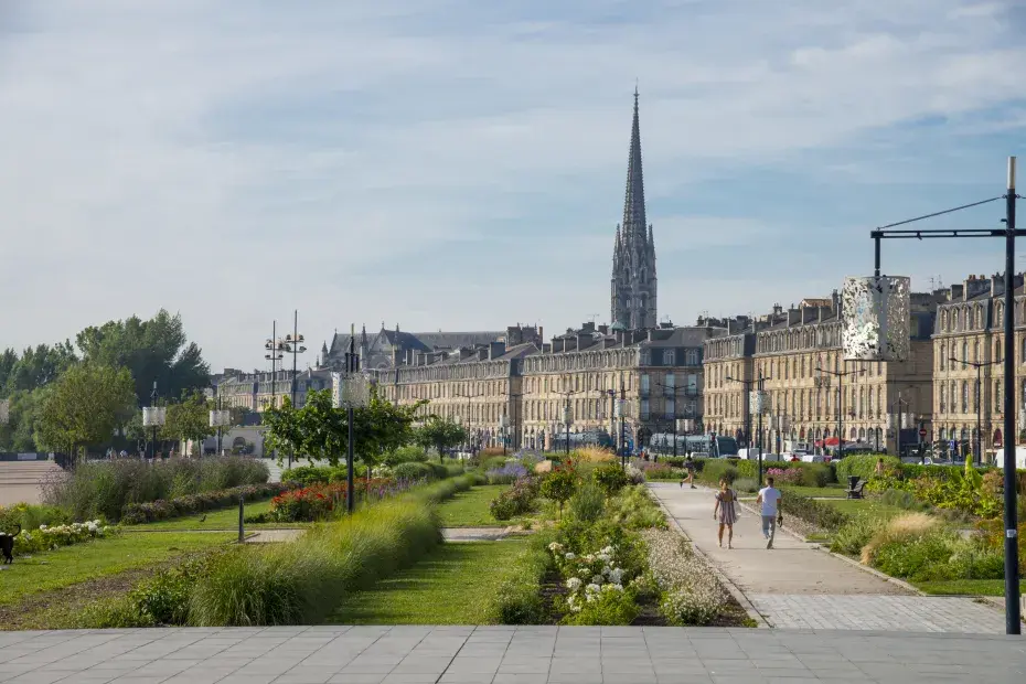 Photo des quais de Bordeaux, avec sur la gauche le jardin des lumières et des passants. Sur la droite, les façades néo-classique des quais, et en arrière plan la flèche Saint-Michel.