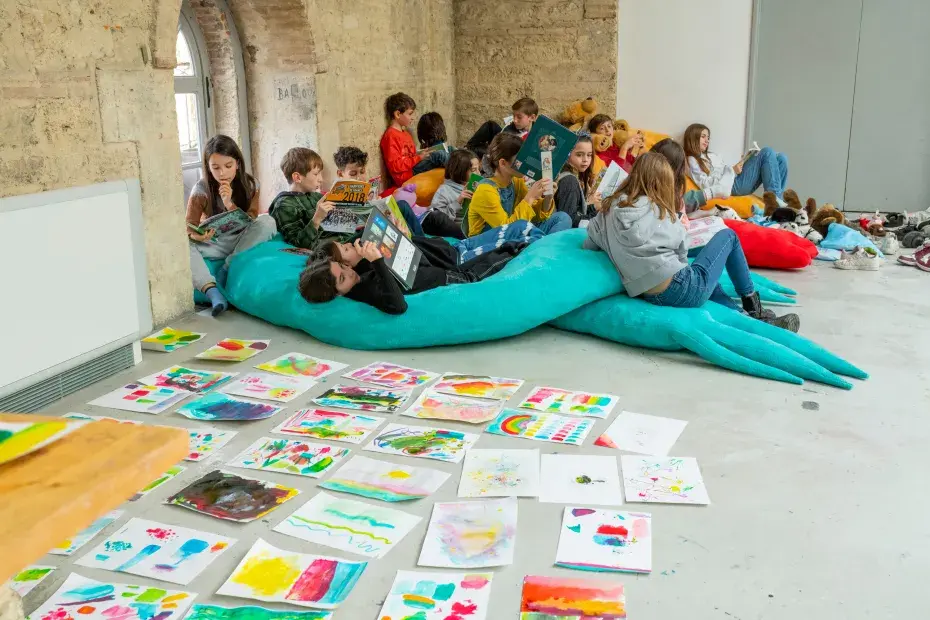 Des enfants lisent et se détendent sur des coussins colorés dans une salle décorée de leurs œuvres artistiques étalées au sol.