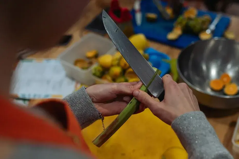Préparation culinaire en cours : une personne épluche une tige végétale à l’aide d’un couteau, entourée d’ingrédients frais, d’ustensiles et de bols sur un plan de travail.