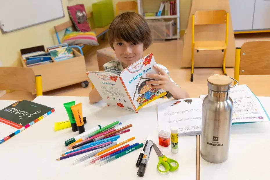 Photo d'un enfant lisant un livre, entouré de fournitures scolaires sur une table.