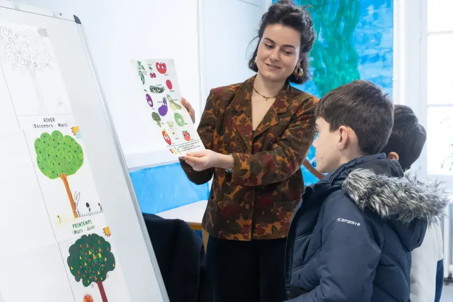 Une diététicienne présente une feuille avec des images de fruits et légumes à deux enfants. Des dessins d'arbres à différentes saisons sont affichés sur un tableau.