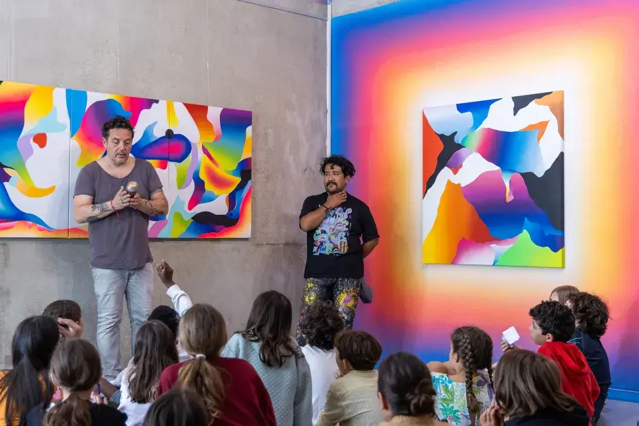 Photo de Pierre Lecaroz et Xomatok devant des enfants assis par terre. En arrière plan, les peintures de Xomatok sont exposées sur les murs