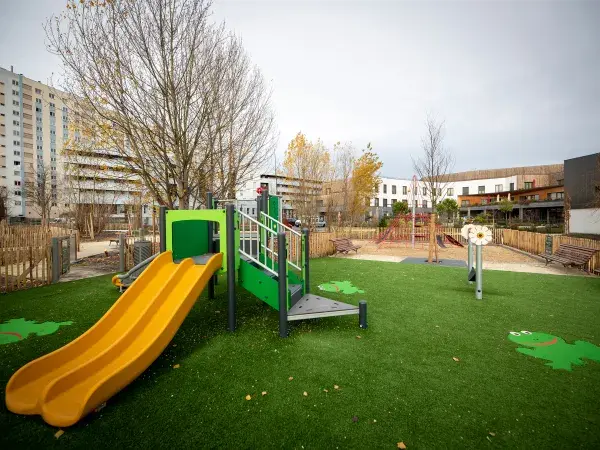 Une aire de jeux colorée avec un toboggan, du gazon synthétique et des immeubles en arrière-plan.