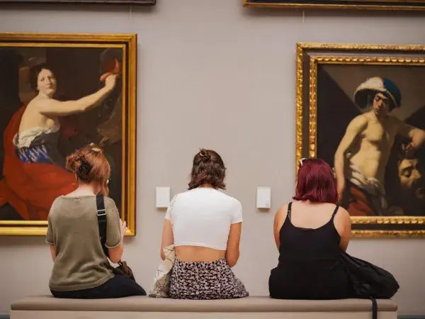 Trois femmes de dos assises sur un banc regardant deux tableaux.