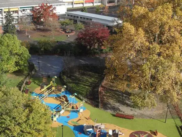 C’est une vue d’en haut d’un parc avec des jeux colorés pour enfants, des arbres et des bâtiments en arrière-plan.
