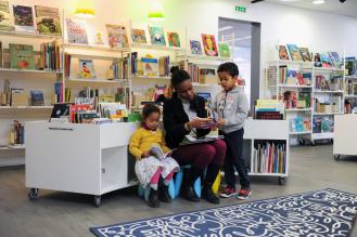 Une femme lit avec deux enfants dans une bibliothèque entourée d’étagères remplies de livres.