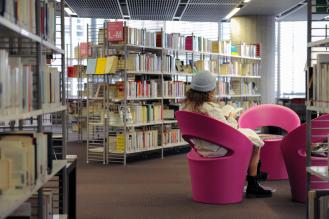 Une personne lit tranquillement dans un fauteuil rose au milieu des rayonnages d'une bibliothèque moderne.