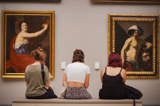Trois femmes de dos assises sur un banc regardant deux tableaux.