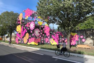 Photo de l'installation "Au delà des boulevards": une structure avec des dessins roses et jaunes, et une cycliste, sur les boulevards