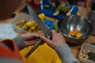 Préparation culinaire en cours : une personne épluche une tige végétale à l’aide d’un couteau, entourée d’ingrédients frais, d’ustensiles et de bols sur un plan de travail.