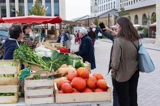 Scène de marché en plein air : des personnes discutent avec un vendeur de fruits et légumes, devant des cagettes remplies de courges, salades, choux et autres produits frais