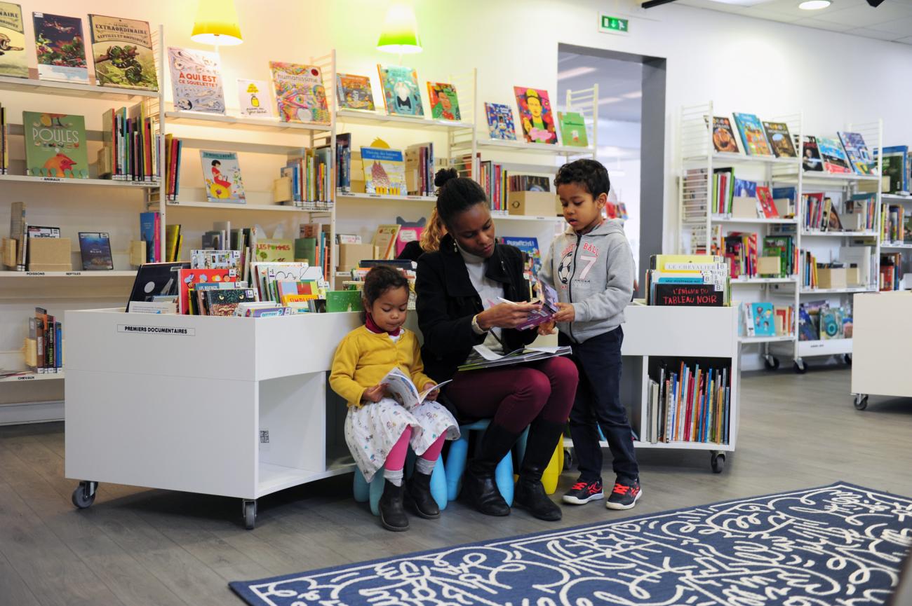 Une femme lit avec deux enfants dans une bibliothèque entourée d’étagères remplies de livres.