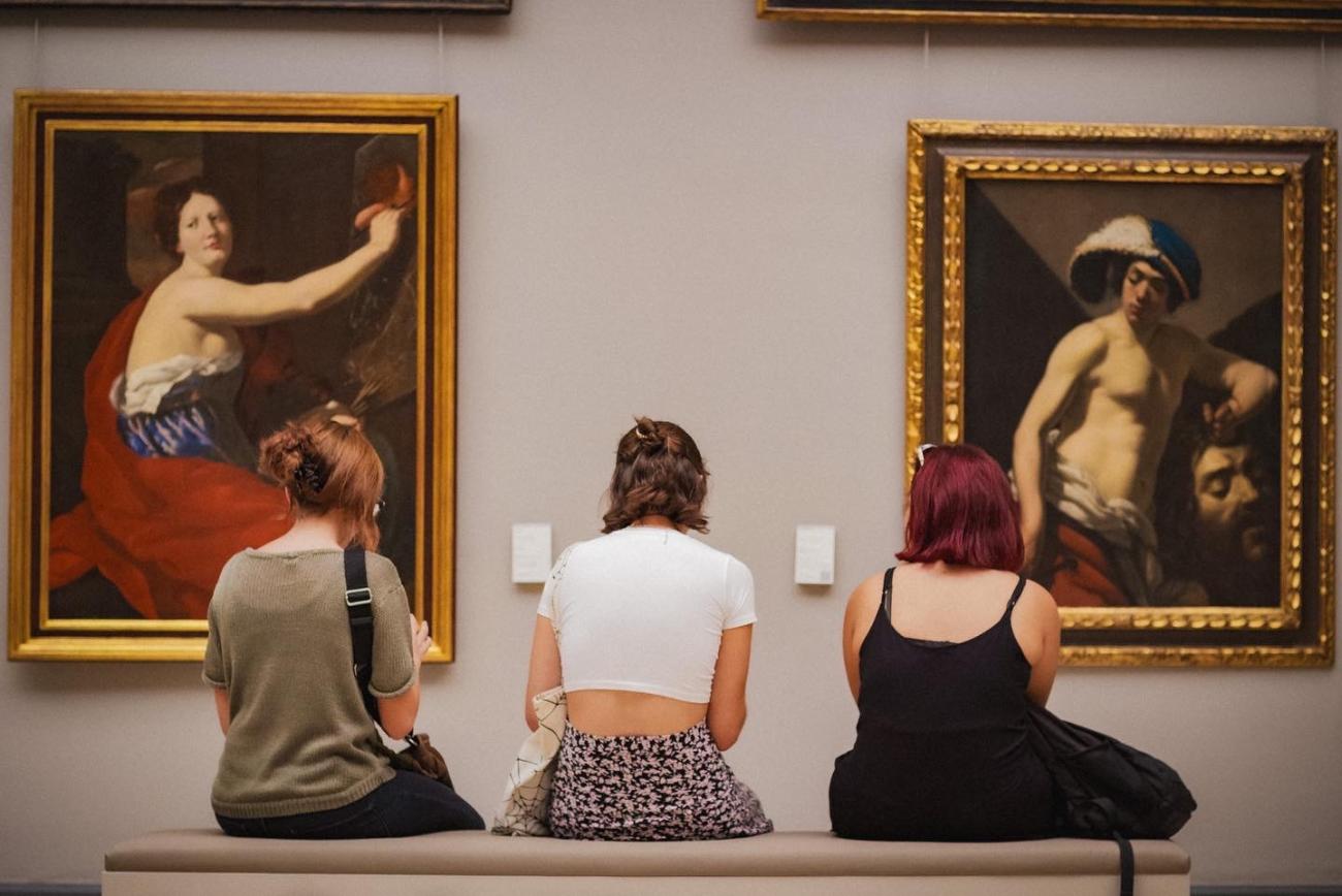 Trois femmes de dos assises sur un banc regardant deux tableaux.